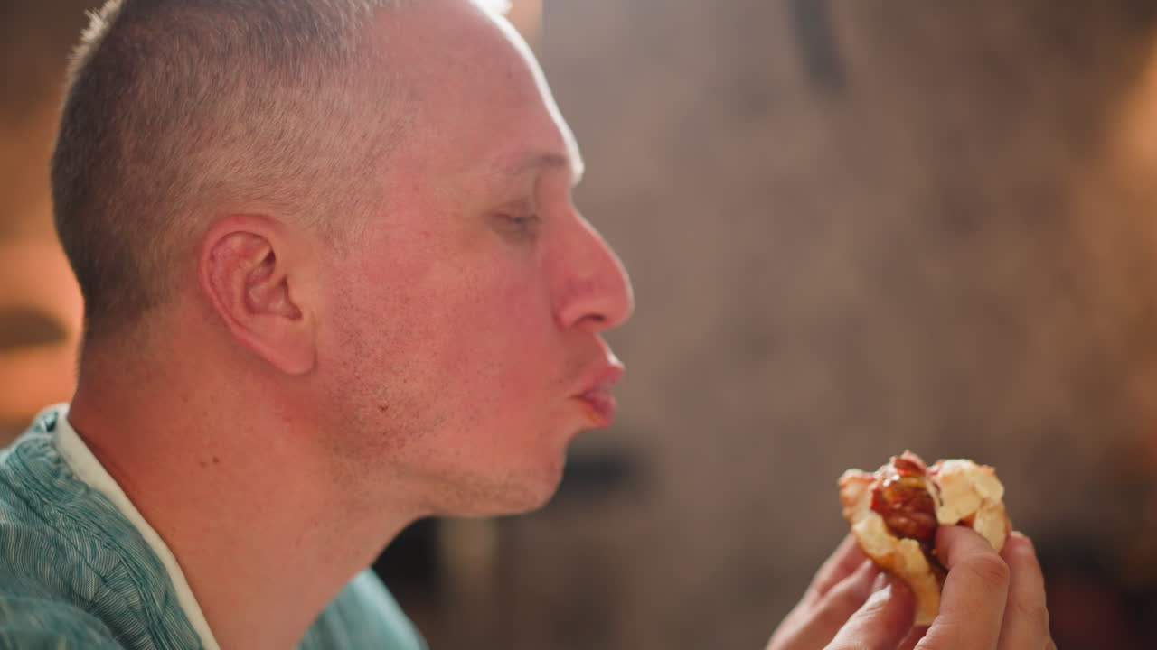 Close side view of adult man eating partially bitten sausage snack indoors with warm light reflecting on face, holding food gently in both hands, cozy dining scene with soft blur background