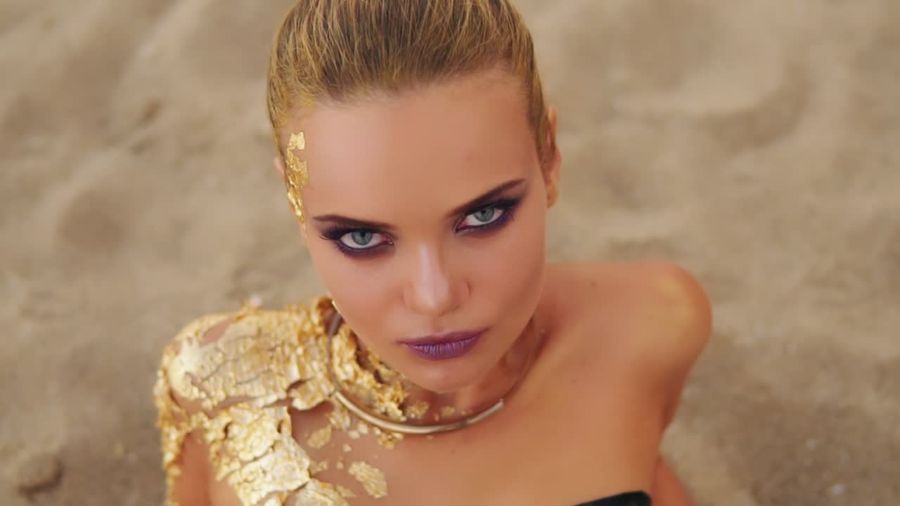 전문적인 골든 아트 메이크업을 한 젊은 모델이 해변에 누워서 카메라를 쳐다보고 있습니다. 매력적인 모델