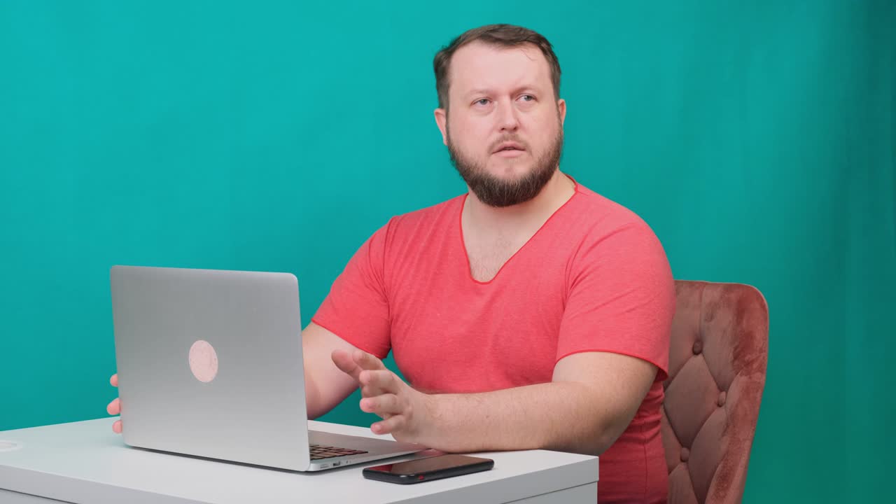 joven empresario feliz con una camiseta rosa trabaja en una computadora portátil y habla con la cámara en una pantalla verde. retrato de un hombre que habla mirando a su computadora portable. hombre trabajando en su escritorio en la oficina