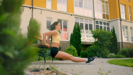 A-young-woman-with-headphones-performs-push-ups-on-a-bench-in-a-city-park-in-slow-motion.-Training-a-young-woman-on-a-bench-against-the-backdrop-of-the-city-and-houses