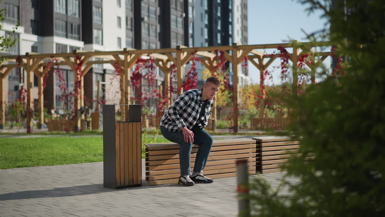 Young man in checkered shirt prepares to sit on wooden bench while reaching into front pocket to get medicine in peaceful outdoor space surrounded by modern buildings, greenery, and soft sunlight