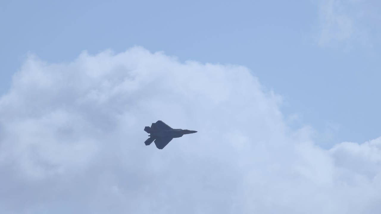 An F-22 Raptor jet flies against a backdrop of clouds, showcasing its agility and power in a clear blue sky