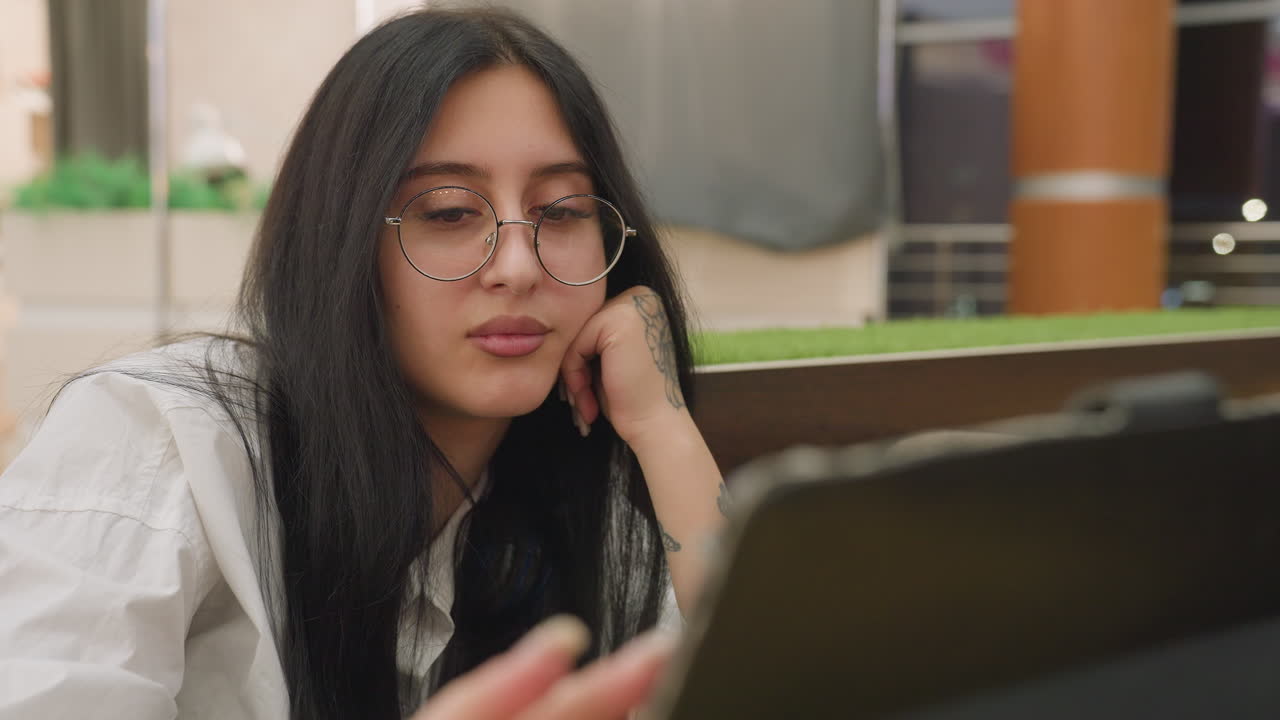Close up of young lady with glasses resting tattooed hand under jaw while operating tablet in well lit mall, eyes closed in relaxed posture, surrounded by soft indoor lighting