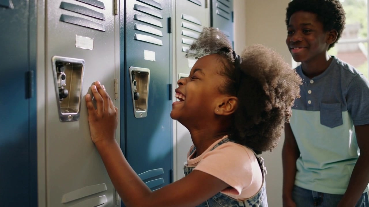 Reaching girl in overalls pressing blue locker dial in school hall, laughing while playing with boy