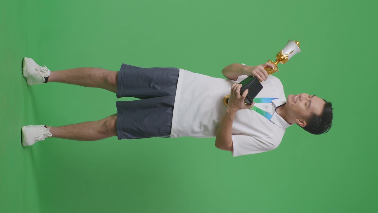 cuerpo lleno de hombre asiático con una medalla de oro besando un trofeo de oro estando orgulloso de ganar como el primer ganador en el fondo de la pantalla verde en el estudio