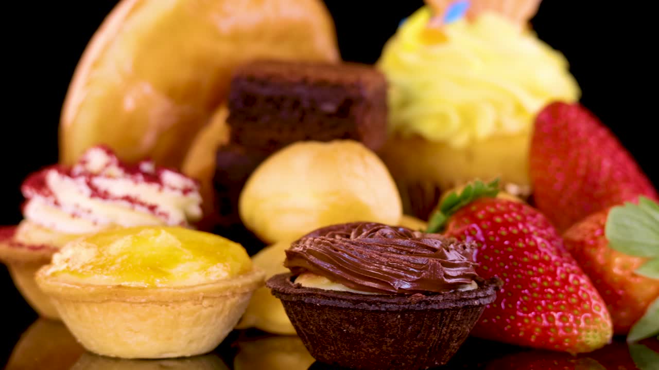 A colorful assortment of cupcakes, donuts, brownies, tarts, and strawberries rotates under bright studio lighting against a seamless black backdrop, highlighting glossy textures and vibrant colors