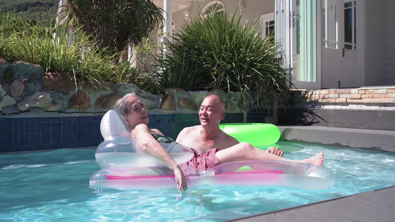 Senior couple chatting, relaxing, adjusting on pink float at backyard pool, small waves nudging it