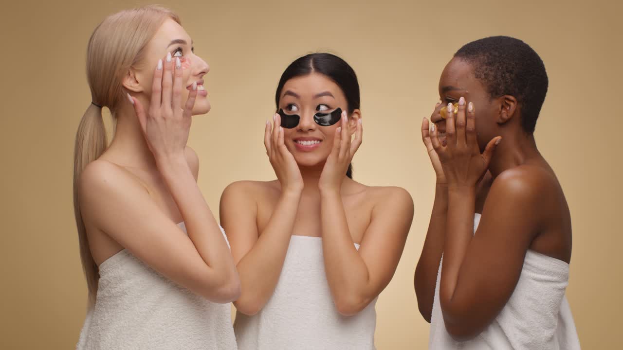 Positive diverse women wrapped in towels putting gel patches on under eyes area and laughing, beige studio background