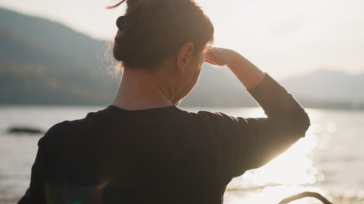 Lady covers face with hand looking ahead. Woman feels lonely sitting on riverbank with scenic view. Gathering thoughts with calm atmosphere of nature