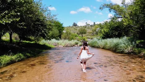 una mujer descalza con un vestido blanco camina alegremente a través de un arroyo de agua dulce en medio del bosque con un dron en cámara lenta