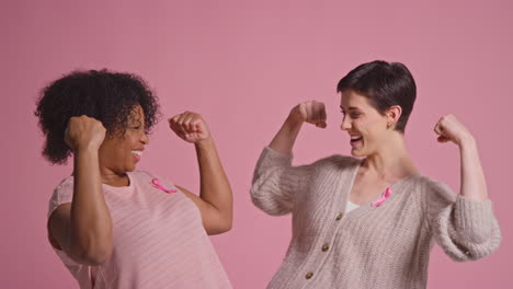 Retrato-De-Estudio-De-Dos-Mujeres-Orgullosas-De-Llevar-Lazos-Rosados-De-Concientización-Sobre-El-Cáncer-De-Mama-Abrazándose-Sobre-Un-Fondo-Rosa