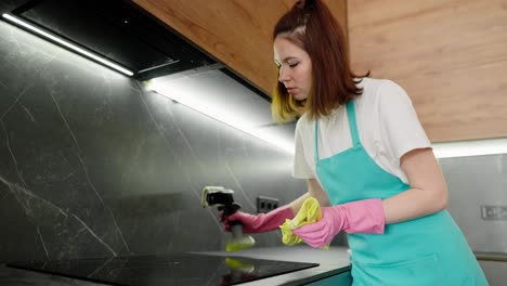 a brunette cleaning lady girl in a white t-shirt and a blue apron uses detergent and a rag to wash a black electronic stove in the kitchen in a modern apartment on a cleaning call