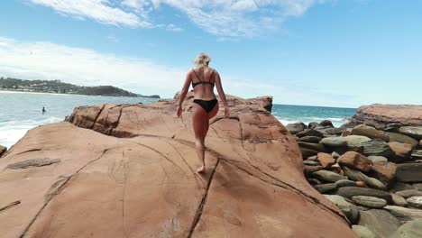 Blonde-girl-walks-on-ocean-rocks-towards-the-ocean-waves-in-slow-motion