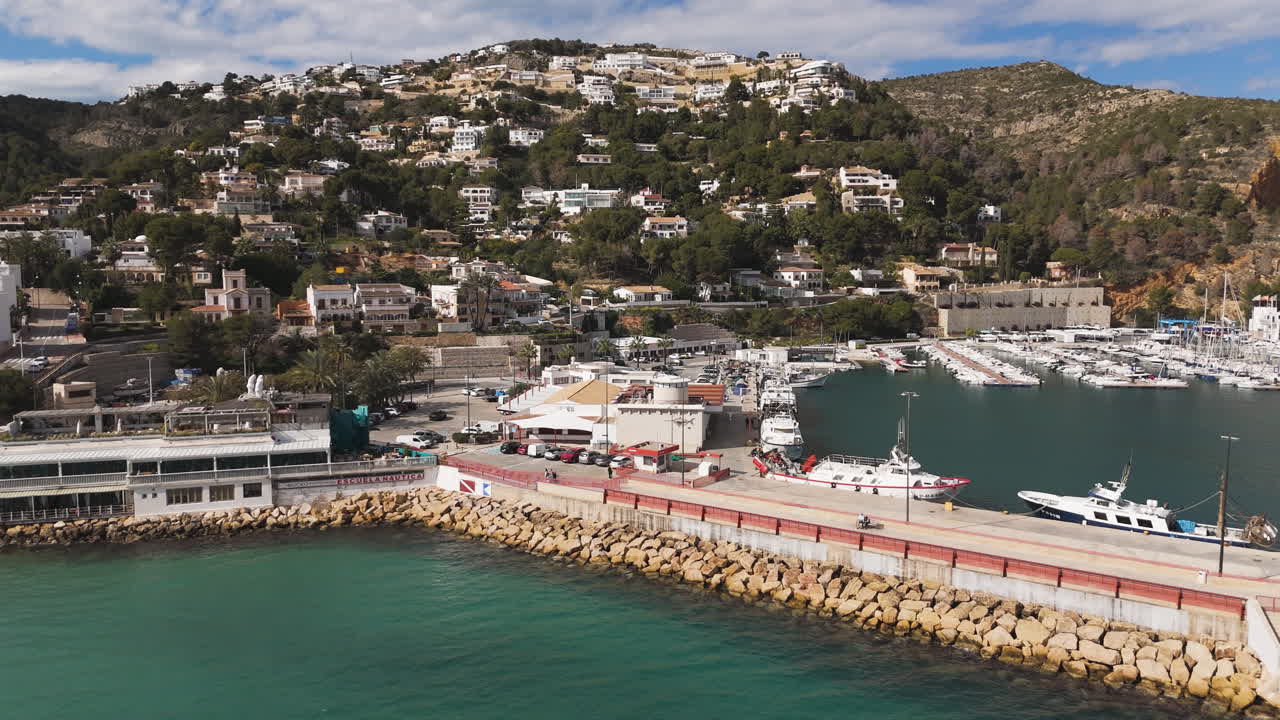 Tilted aerial shows turquoise bay next to marina promenade and distant hillside homes