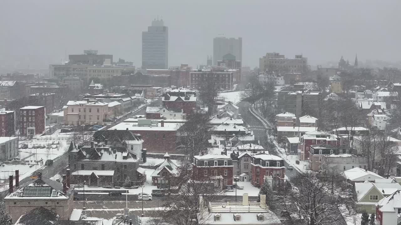 Downtown Worcester skyline with City Hall on the horizon. Brick homes sprawl below. Overhead vantage frames the urban layout, aerial zoom. Snow flurries flying in Massachusetts.