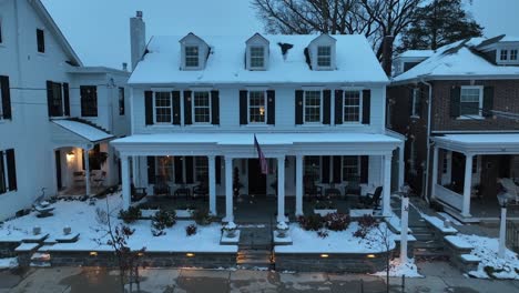 snowy two story building with american flag at front exterior of house. snowflakes falling from sky. family time in christmas season. aerial approaching shot.