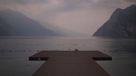 Muelle-De-Madera-En-La-Playa-De-Riva-Del-Garda,-Con-Lago-De-Garda-Y-Montañas-Al-Fondo