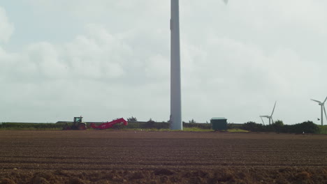 wind turbine spinning while farmer turns soil in a field in cornwall - tilt down wide shot