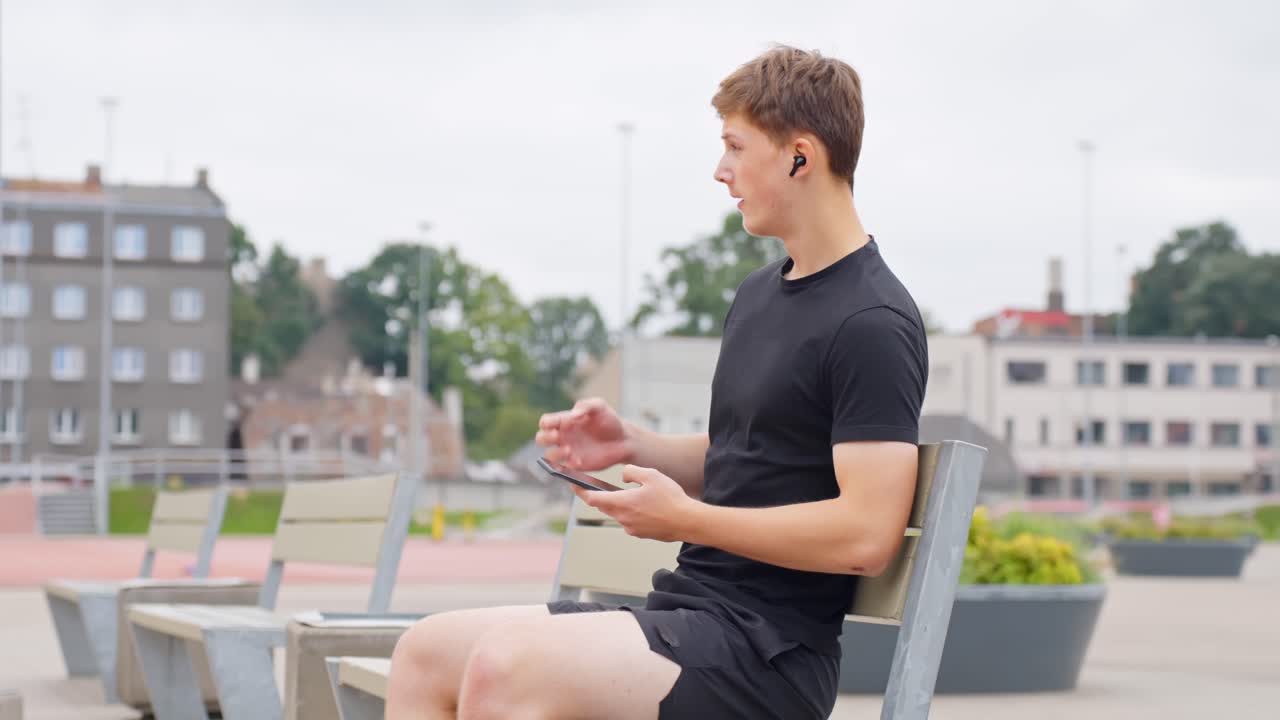Man sits alone on park bench scrolling phone and listening to music, pre-exercise calm
