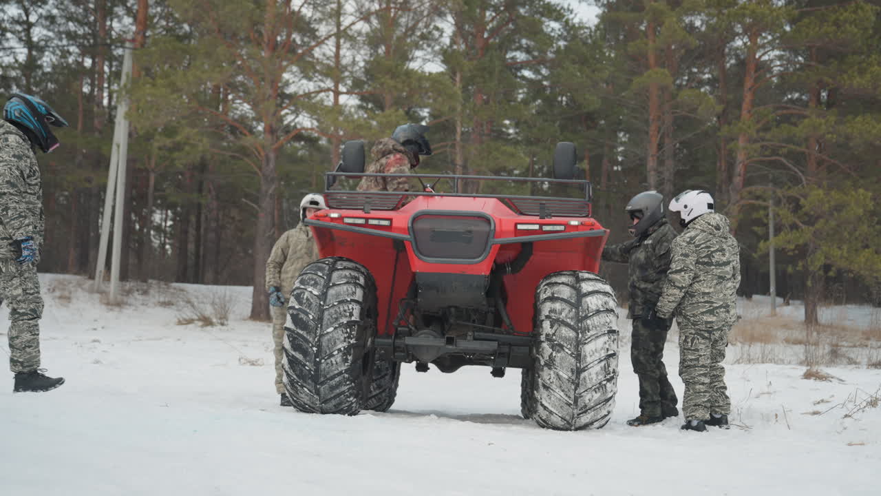 ATV rosso parcheggiato in una foresta innevata, equipaggio che ispeziona pneumatici e verricello, guida che indica il sentiero, pilota che sale per una spedizione fuoristrada, regolazione dell'equipaggiamento, conversazione di avvertimento, attesa prima di un'esperienza estrema.