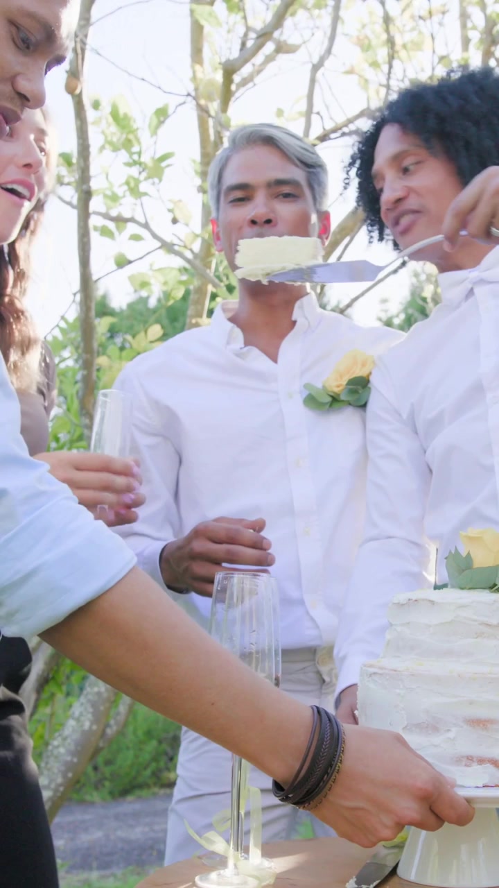 縦型動画：結婚式で白いシャツを着た男性たちが庭でケーキを切り、サーバーに手を伸ばして一切れを配る。