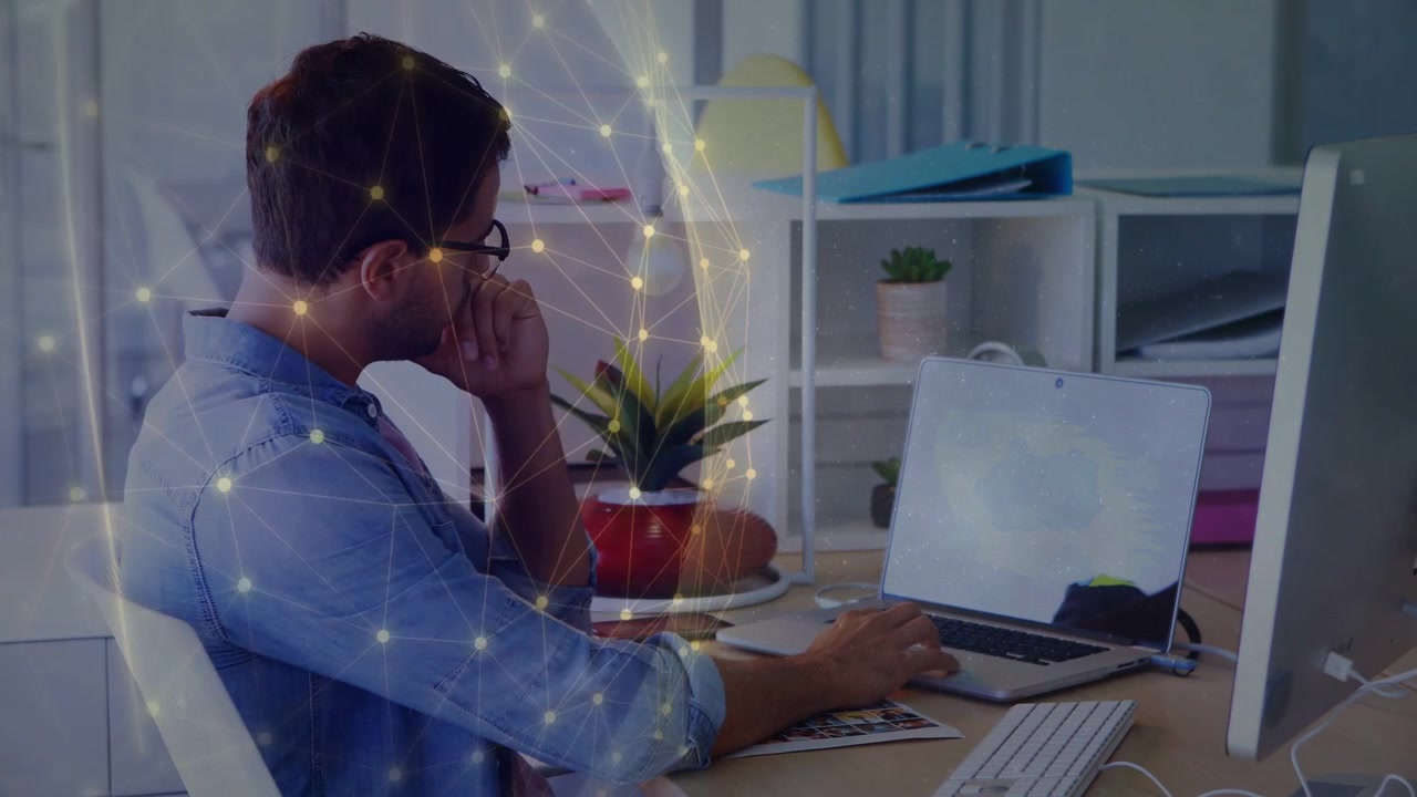 Uomo in camicia blu seduto nell'ufficio di casa che digita sulla tastiera, usando un laptop, un monitor e nodi dorati