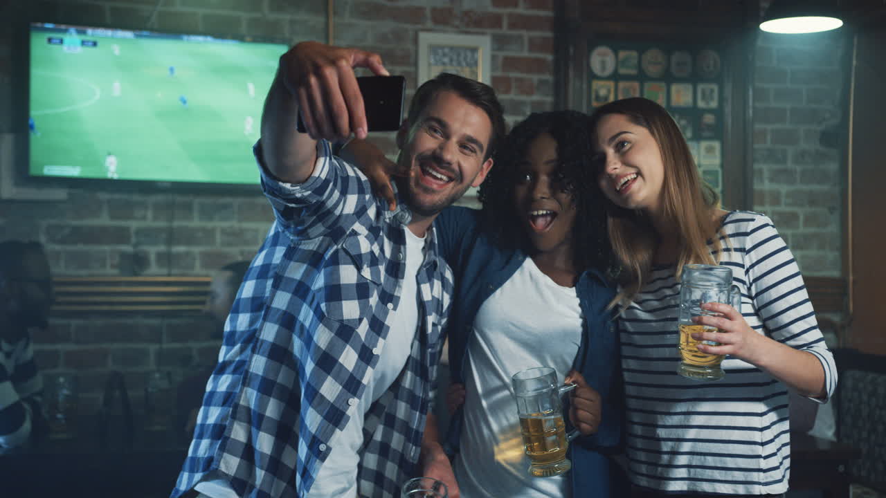 jóvenes amigos multiétnicos en el pub durante el juego deportivo, hombre tomando una foto selfie en el smartphone con dos mujeres