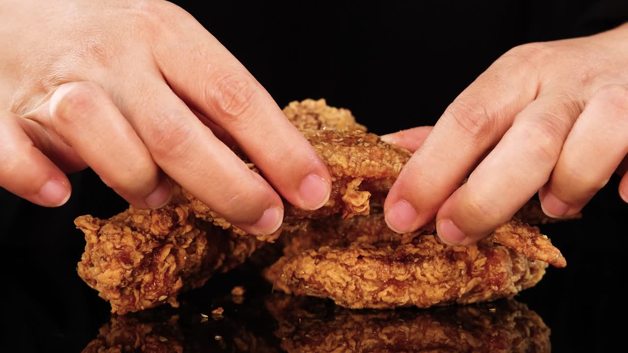 Close-up sequence of hands tearing apart a crispy fried chicken wing on a glossy black surface, under bright, even lighting with minimal camera movement