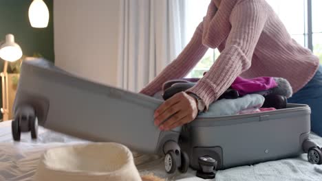 midsection of biracial woman packing suitcase in sunny bedroom, slow motion