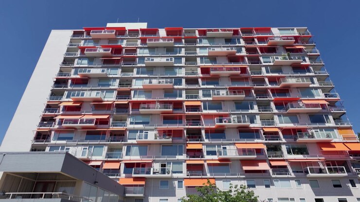 Plano estático amplo de um edifício residencial moderno de vários andares com inúmeras varandas e toldos vermelhos. Um edifício cinza mais baixo e folhagem verde aparecem em primeiro plano, emoldurando a cena urbana.