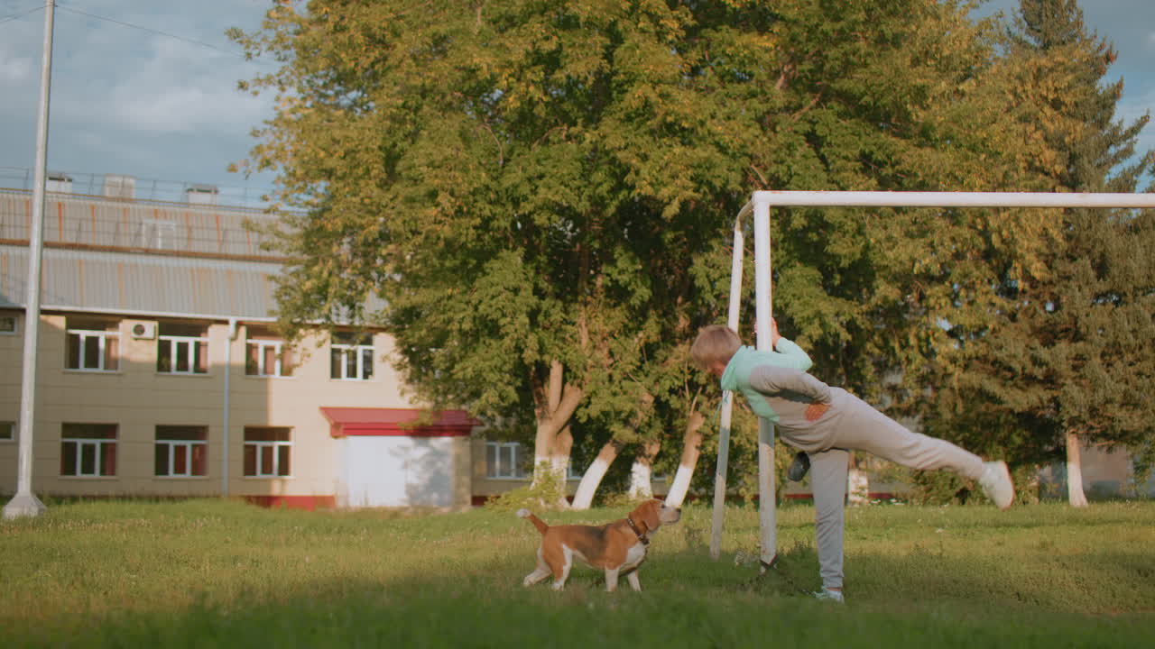 Trainer holding football post performing knee to chest stretch during sunset while german shepherd tied nearby on grassy field with trees and building in background showcasing fitness