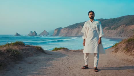 indian man wearing traditional white clothes is posing on a cliff overlooking a beautiful ocean with waves crashing on the shore and mountains in the background during a sunny day