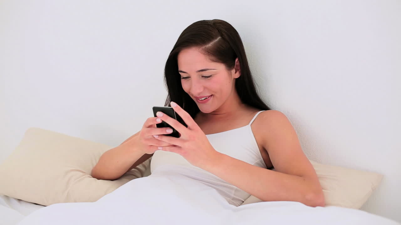 매력적인 갈색 여성이 침대에서 스마트폰으로 문자 메시지를 보내고 있습니다.