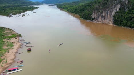 Dschunke-Bringt-Besucher-über-Den-Mekong-Zu-Den-Pak-Ou-Höhlen,-Während-Eine-Drohne-Herabsinkt