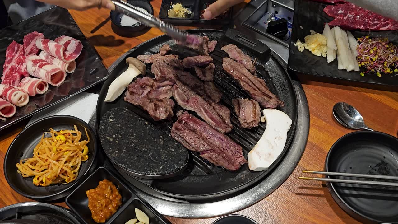 Close up shot of a person using scissors to cut sizzling grilled beef and galbi ribs on a traditional tabletop Korean barbecue surrounded by delicious banchan