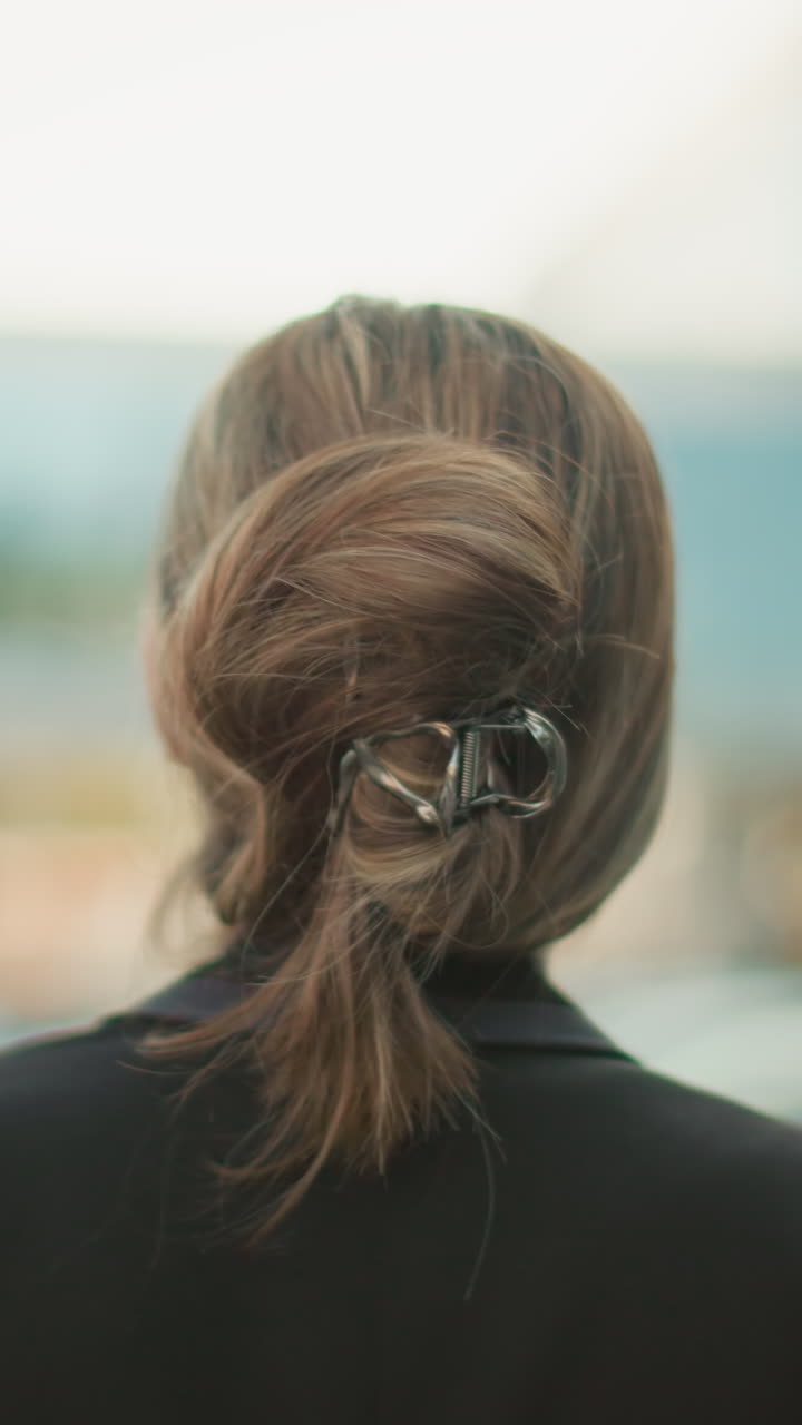 Close up back view of woman with neatly clipped hair glancing left with blurred background of parked cars and modern glass building in city environment during bright workday morning in urban setting