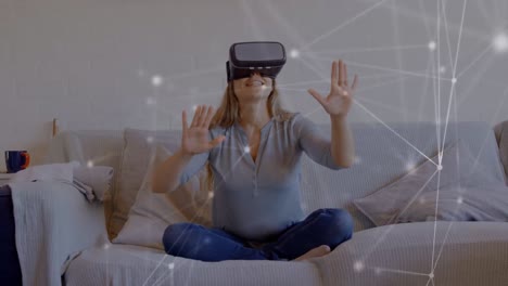 Network-of-connections-over-caucasian-woman-wearing-vr-headset-sitting-on-the-couch-at-home