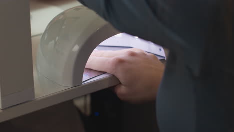 close-up of customer placing hand inside led nail dryer to cure freshly applied polish during manicure session at salon, showing careful positioning under light for proper drying process