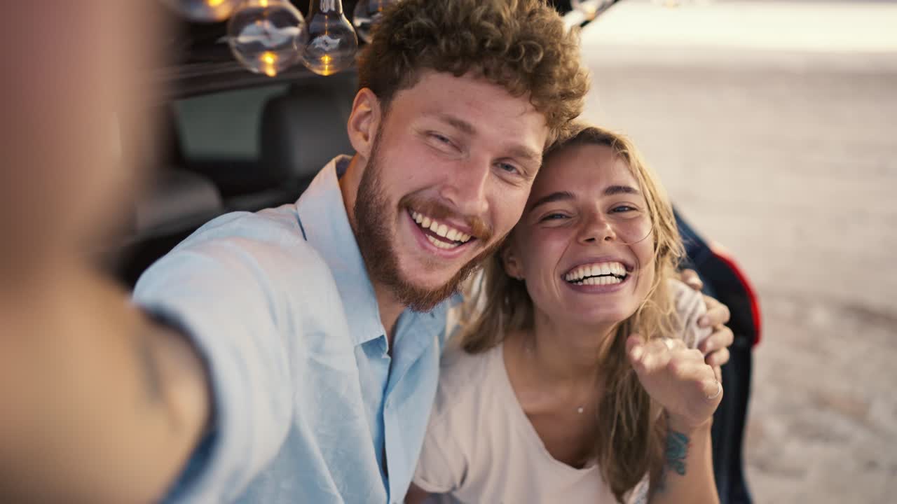 pov: un tipo barbudo con cabello rizado en una camisa azul está sonriendo a la cámara con su novia rubia mientras agita toma un
