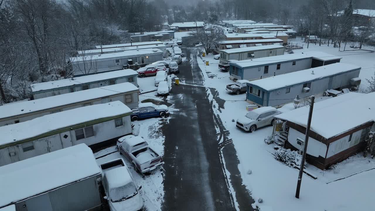 Parque de casas móviles cubierto de nieve en Estados Unidos, con carreteras heladas, coches aparcados y bosque invernal. Frío barrio rural estadounidense en temporada navideña. Toma aérea hacia atrás