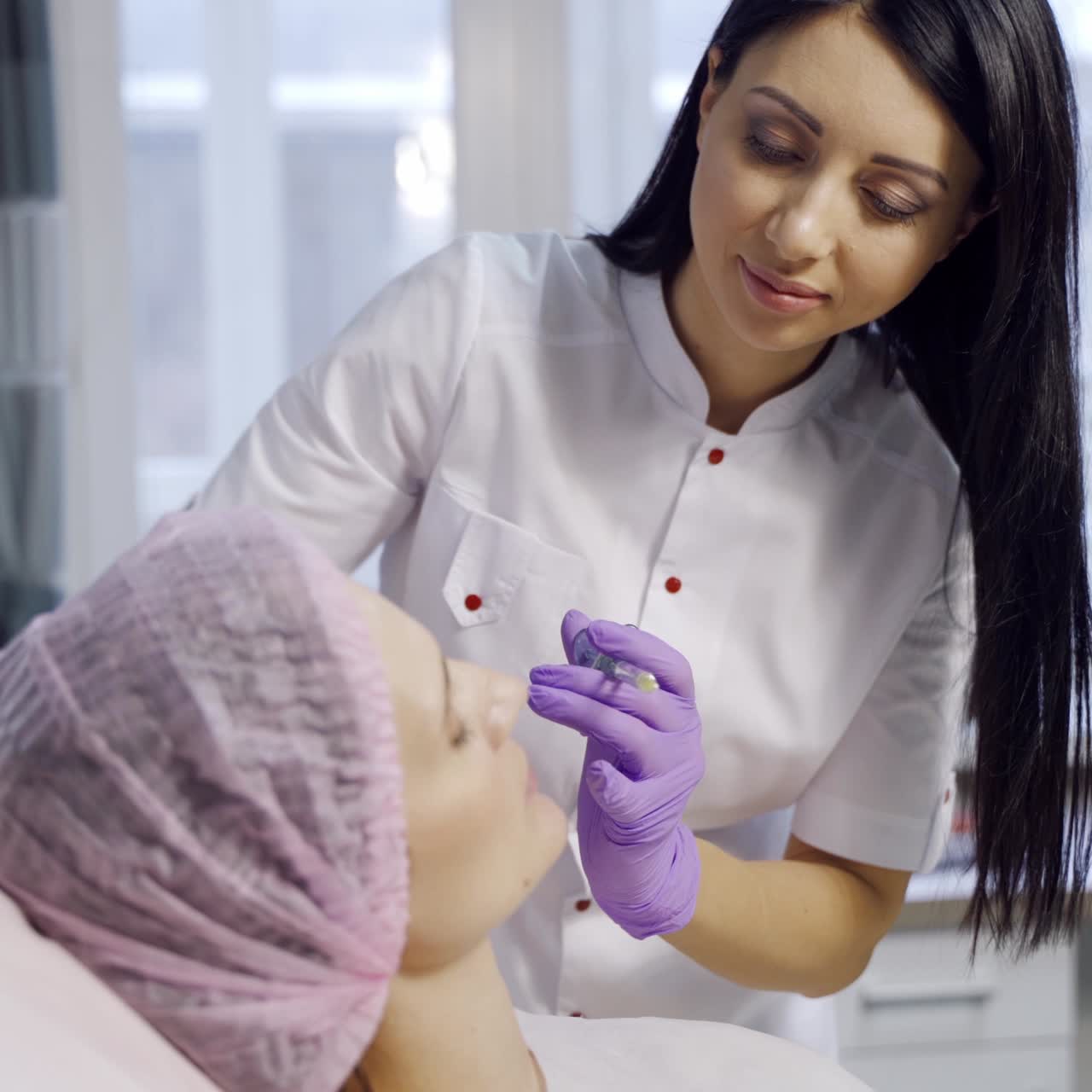 A female cosmetologist makes beauty injections for a client that lays on the couch in the room near a window in the center of cosmetology. Beauty industry.