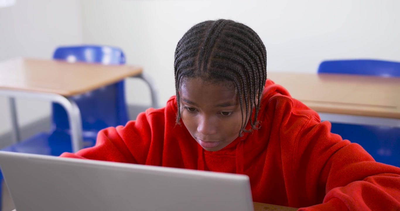 Adolescente afroamericano con felpa rossa che inizia un compito sul laptop, chinato a digitare al banco di scuola.