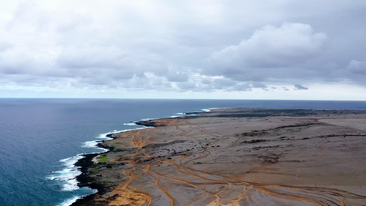 Wide view of a volcanic coastline stretching into the distance with rugged dirt tracks, crashing waves, and a dramatic overcast sky along the Pacific Ocean.