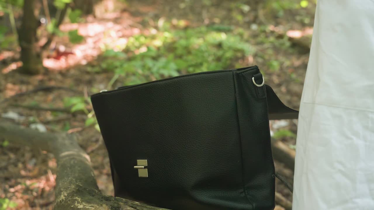 Close view of black backpack placed on old tree stump as hand reaches to open clasp preparing to bring out jotter and pen in natural outdoor setting with blurred background of forest ground