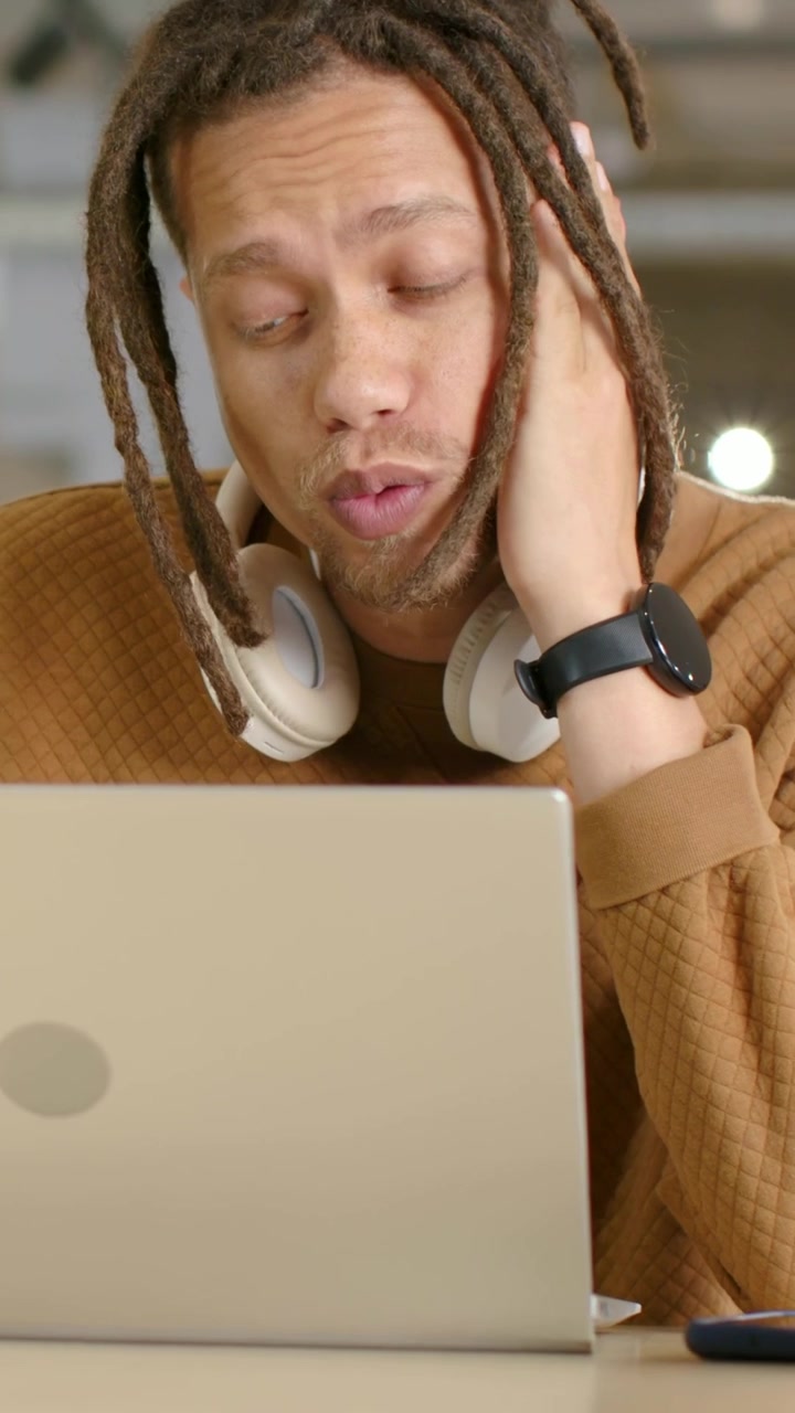 Vertical video: Leaning man in brown sweater processing laptop content at table, headphones, watch