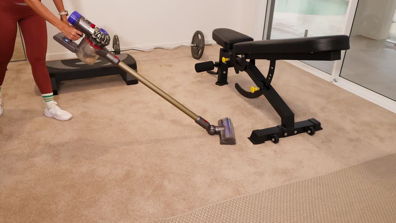 A woman vacuums the carpet in a well-lit home gym, surrounded by fitness equipment, creating a clean and organized environment