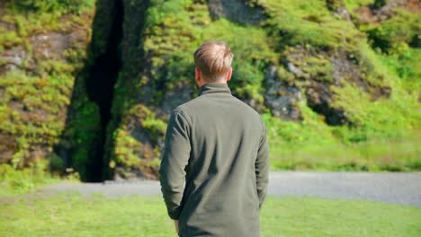 back of the man walking on the green meadow towards the cave of seljalandsfoss waterfall in iceland