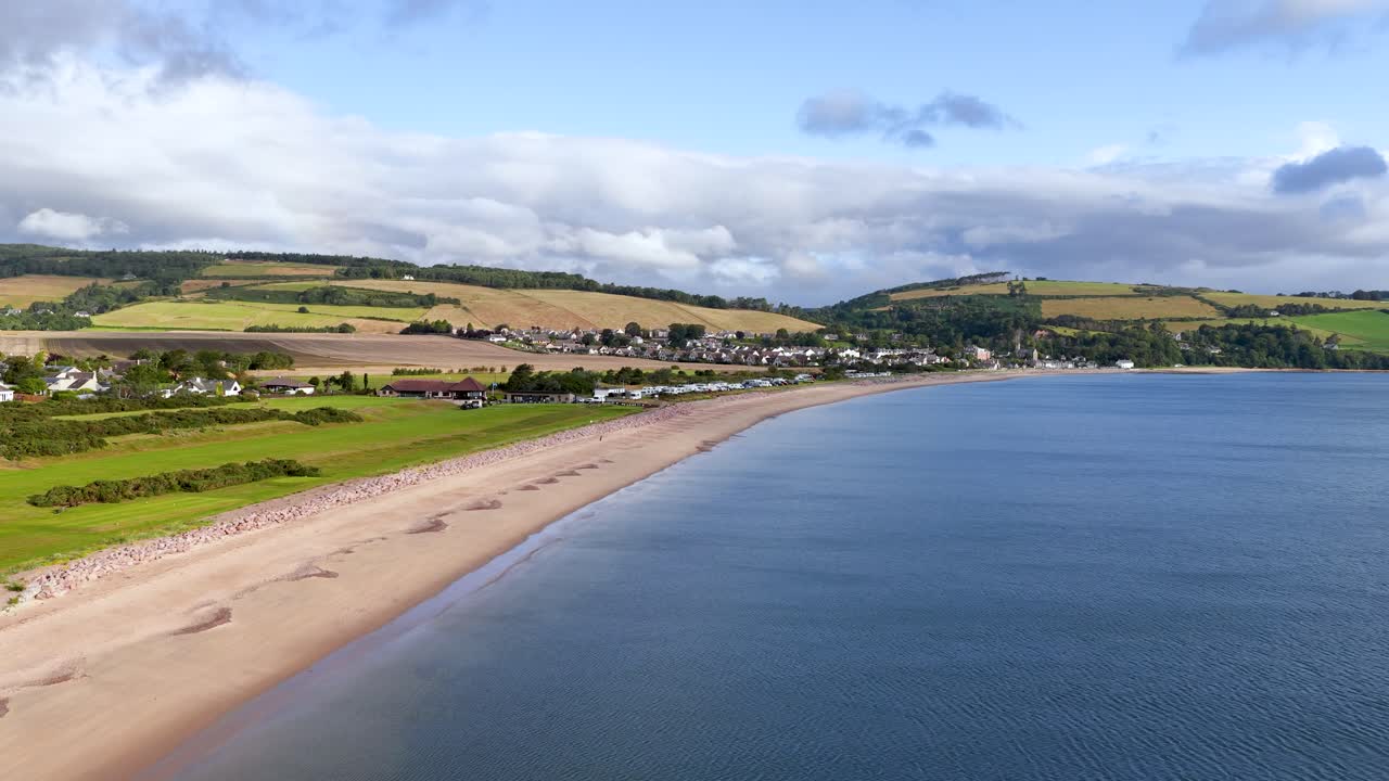 Drone glides above tranquil Rosemarkie shoreline, revealing village, hills, and calm summer waters