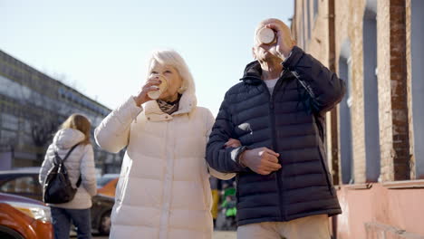 vista frontal de una pareja mayor hablando y caminando por la calle en un día de invierno mientras toma cafés para llevar