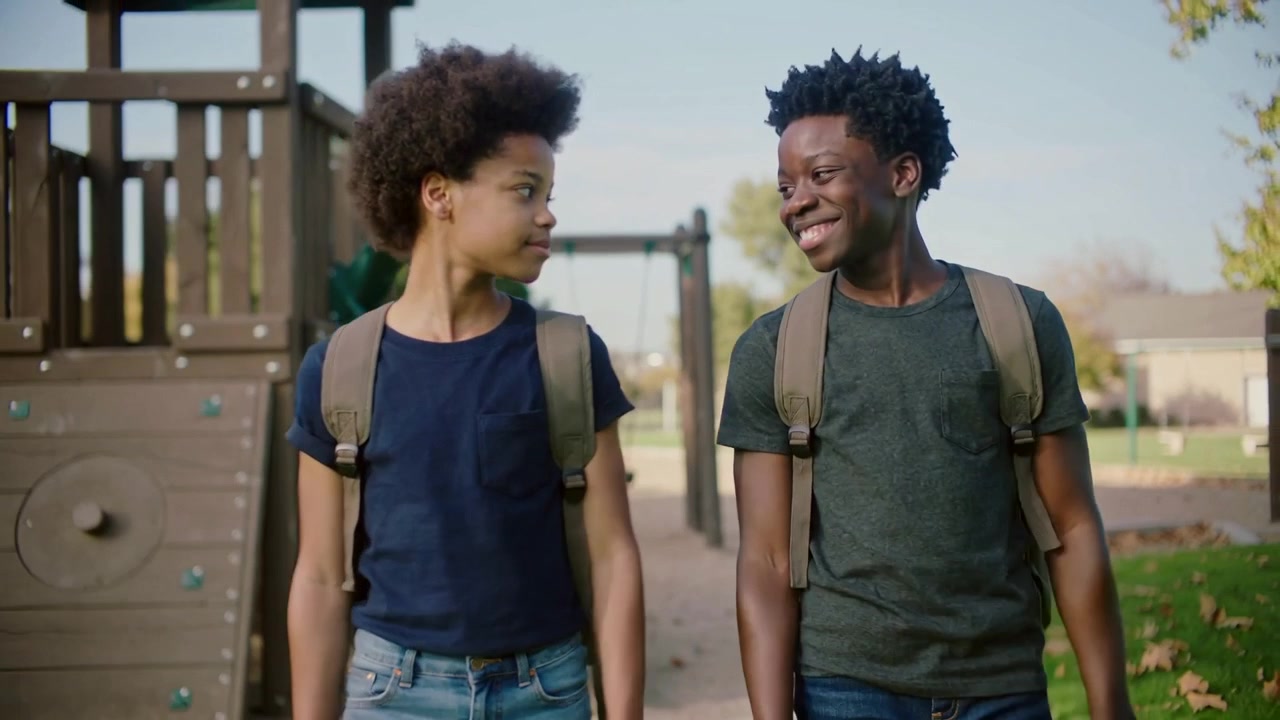 Exchanging glance, two teens walking in park wearing short sleeves with tan backpacks after school